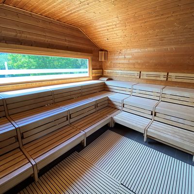 Dieses Bild wurde in der Außensauna aufgenommen, Sie sehen eine große Eckholzbank mit drei Sitz-/ Liegeebenen. Aus dieser Sauna können Sie über ein Panoramafenster nach draußen ins Grüne schauen.